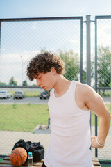Naklejka premium A young man with curly hair adjusts his tank top while standing on an outdoor basketball court. The bright sunlight illuminates the area, creating a vibrant atmosphere.