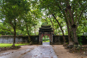 Obraz premium A historic, ornate stone gate stands between leafy trees and a stone wall at Minh Mang tomb, Hue.