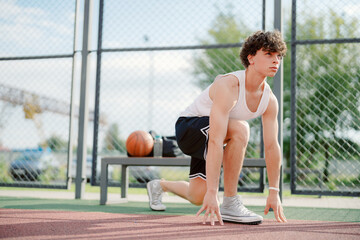 A young male athlete is crouched in a ready position on an outdoor basketball court. The sun is shining, and a basketball rests nearby, emphasizing his focus on the upcoming drill.