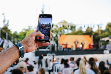 hand holding a smartphone to take a photo or record a live performance or concert with a blurred background of stage lights and bokeh