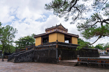 The Stele Pavilion, a two-tiered, yellow-painted structure with a tiered platform and stone staircase, stands under a cloudy sky at Minh Mang tomb, Hue.