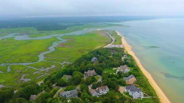 Aerial view of expansive coastal marshes meeting the sandy shoreline on an overcast day, with residential beach houses nestled among trees and calm ocean waters stretching into the distance.