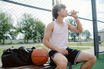 A young male athlete sits on a bench, enjoying a refreshing drink after a basketball practice. The setting is a sunny park designed for sports, surrounded by trees and open space.
