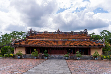 Fototapeta premium The Minh Lau pavilion, a long, single-story building with a red-tiled roof and dragon ornaments, stands centered on a brick courtyard under a cloudy sky at Minh Mang tomb, Hue.