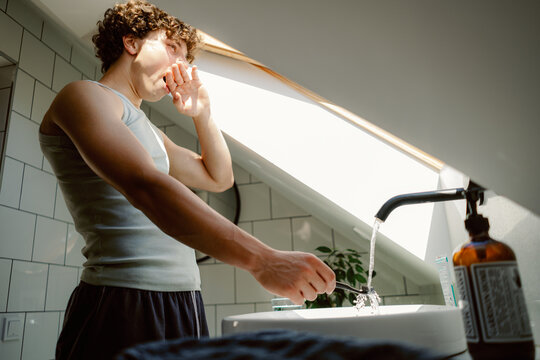 A young man stands at a bathroom sink, brushing his teeth while yawning. Sunlight streams through a window, illuminating the space and creating a fresh morning atmosphere.