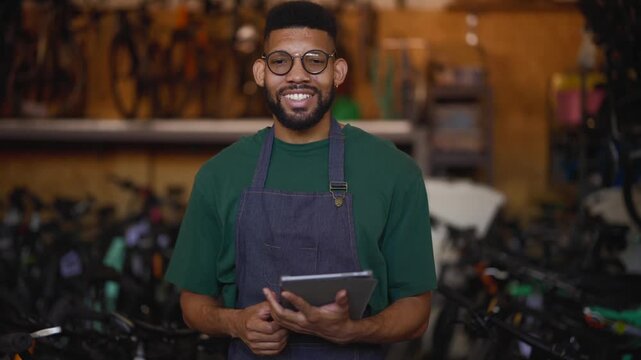Friendly bicycle mechanic wearing protective apron and glasses, holding digital tablet and smiling confidently inside modern bicycle repair workshop with blurred background