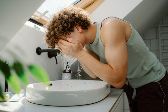A young man with curly hair is washing his face in a modern bathroom. Sunlight streams through a skylight, highlighting the clean design and natural elements. - Powered by Adobe