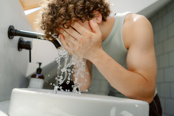 A young man with curly hair is washing his face at a stylish bathroom sink. Sunlight streams in through a window, illuminating the scene and water splashes around him.
