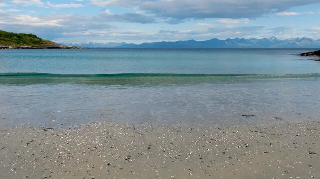 Experience the serene beauty of Mannen Haukland beach in Lofoten, Norway, where gentle waves lap at the sandy shore. The picturesque landscape showcases distant mountains and clear blue skies.