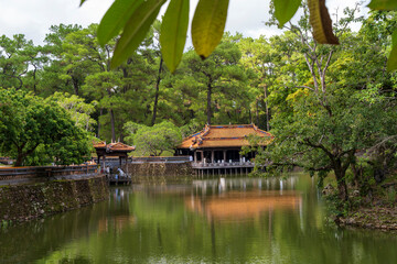 A tranquil lake reflects the Hien Khiem Pavilion and surrounding lush pine forest under a cloudy sky at Tu Duc tomb, Hue.