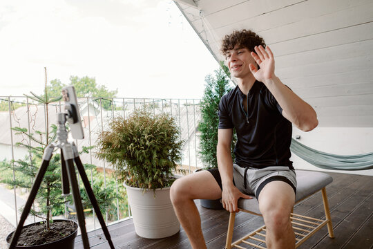 A young man with curly hair sits on a stool, waving while engaging in an online video call from a bright balcony surrounded by plants on a sunny day.