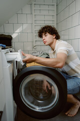 A young man with curly hair is crouching beside a washing machine in a well-lit laundry area. He is focused on loading laundry while surrounded by white tiles and natural light.