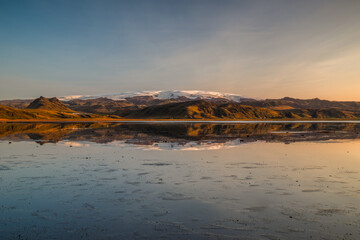 Golden evening light bathes the rugged Icelandic landscape, with the ice cap of the glacier Mýrdalsjökull reflecting in shallow waters of a black sand beach