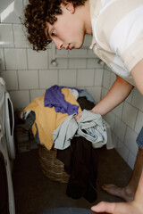A young man sorts his clothes as he prepares to do laundry at home. He stands near a washing machine, focusing on organizing his garments in a cozy laundry area.