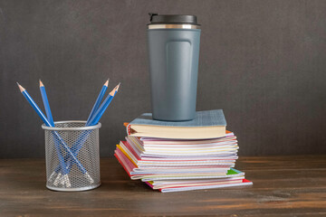Back to college concept with thermos cup, stack of notebooks on dark desk against the background.