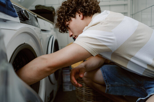A young man with curly hair is focused on loading clothes into a washing machine. The room is bright and airy, and various laundry items are scattered around.