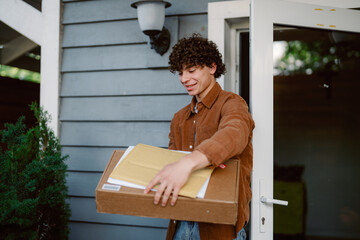 A young man with curly hair is happily accepting a package at his front door. He is dressed casually and standing in a welcoming home environment during daylight.