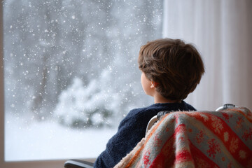 Young boy in wheelchair with blanket looks out window at peaceful, snowy winter scene. child sits with quiet contemplation, watching falling snow outside