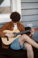 A young man with curly hair is sitting on a patio, focused on playing his acoustic guitar. He wears a brown jacket and denim shorts, enjoying a relaxed afternoon outdoors.