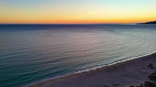 A beautiful sunset over the ocean with gentle waves rolling onto a sandy beach beside a large hill.