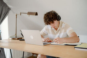 A young student sits at a wooden desk, focused on his laptop while wearing headphones. Books and notebooks are nearby, indicating a study session in a cozy space.