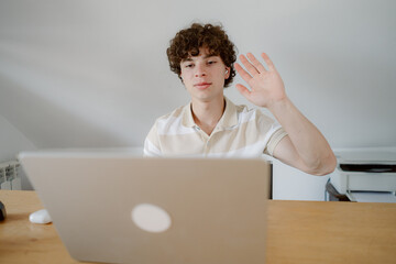 A young man with curly hair sits at a wooden desk, waving at his laptop while engaging in a virtual meeting in a bright home office. Sunlight filters through the room.