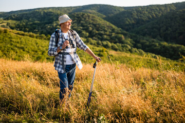 Happy senior man with backpack and hiking poles using binoculars while hiking in nature.	