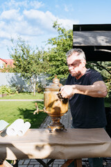 An elderly man carries a traditional brass samovar heated outdoors in a green garden into the house.