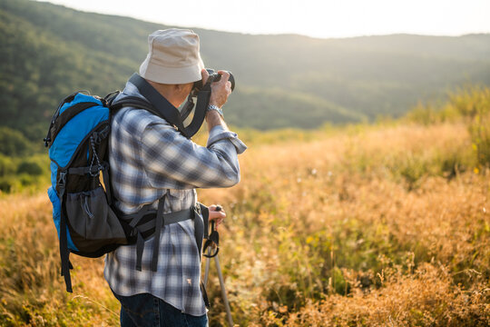Active senior hiker using camera for photographing while hiking with backpack and hiking poles in nature.
