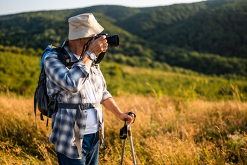 Active senior hiker using camera for photographing while hiking with backpack and hiking poles in nature.