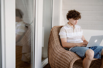 A young man sits comfortably on a wicker chair on a balcony, focusing on his laptop. The sunlit space has a relaxed atmosphere, perfect for outdoor work.