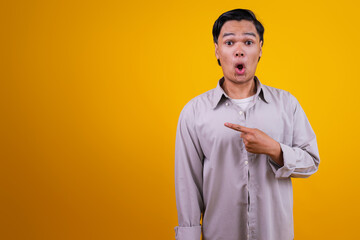 Surprised Young Asian Male Pointing To Side With Copy Space Isolated On Yellow Background