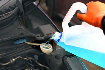 Hand in glove pouring into car reservoir tank blue antifreeze washer liquid, seasonal winter vehicle care and regular automotive maintenance process. Car deicing liquid refill in winter season