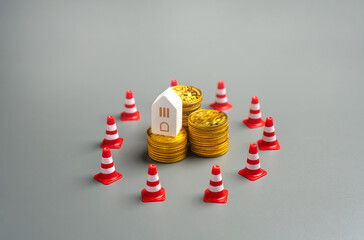 Stacked coins and house surrounded by traffic cones. Asset freeze, financial sanctions, foreclosure risks, and restrictions on property investments due to economic or legal barriers.