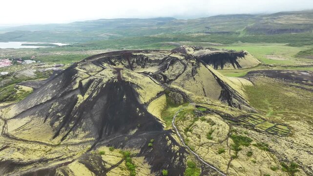 Rocky volcanic grabrok volcano crater during sunny day vegetated with green plants and moss. Lake fjord in distance. Aerial wide shot.