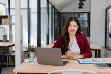 Asian businesswoman is working happily with laptop computer and using technology in modern office, smiling and looking confident, professional worker, business and finance concept