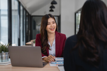 Two Asian businesswomen discussing marketing strategies while analyzing graphs and utilizing a laptop during a meeting in a modern office environment