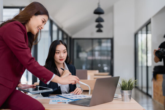 Two asian businesswomen are analyzing financial charts and data on a laptop and tablet, discussing and collaborating on a project in a modern office while a photographer takes pictures of them