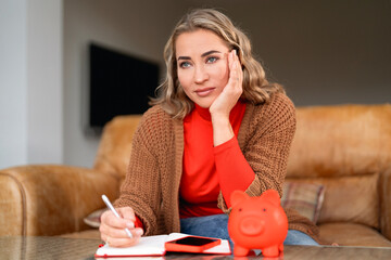 Young woman reflecting on finances while planning budget at home with a piggy bank and notebook on a cozy couch