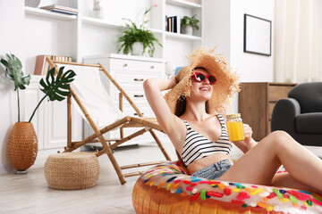 Obraz premium Happy woman in sunglasses and straw hat with mason jar of juice and inflatable ring having fun on floor at home. Dreaming about vacation at sea