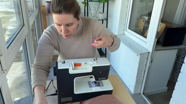 Attractive woman using sewing machine, in addition to scissors, threads, fabric, making clothes at home as little businesswoman