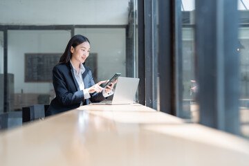 Young Asian businesswoman wearing an elegant suit, working on a laptop and smartphone while sitting by a bright window in a modern office space, exuding professionalism and success