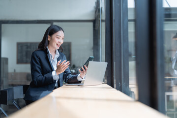 Asian businesswoman using laptop and smartphone having video conference with client from office, she is smiling and gesturing while looking at laptop screen