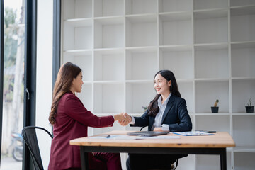 Two asian businesswomen shaking hands after closing a deal sitting at a desk in a modern office, they are smiling and satisfied with the agreement