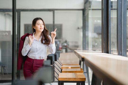 Young asian businesswoman standing in modern office cafeteria is holding jacket on shoulder and pointing finger at window while thinking about future projects