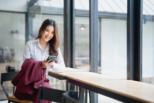 Young Asian businesswoman sitting at a cafe table, smiling while using her smartphone. Enjoying a well-deserved break from work, feeling relaxed and happy in a modern setting