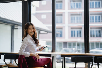 Young Asian businesswoman holding a smartphone and smiling while sitting in a modern office workplace, enjoying work-life balance with technology and connectivity