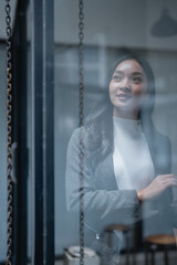 Young businesswoman looking out the office window, smiling with hope and optimism for future success in her career. Contemplating aspirations and envisioning achievements ahead © MINAE