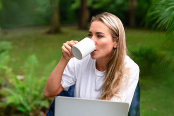 Woman enjoying coffee outdoors while working on a laptop in a serene, green garden setting on a...