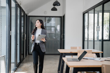 Young Asian businesswoman holding digital tablet walking in modern office and thinking about future projects, startup, new business, company growth, or company development